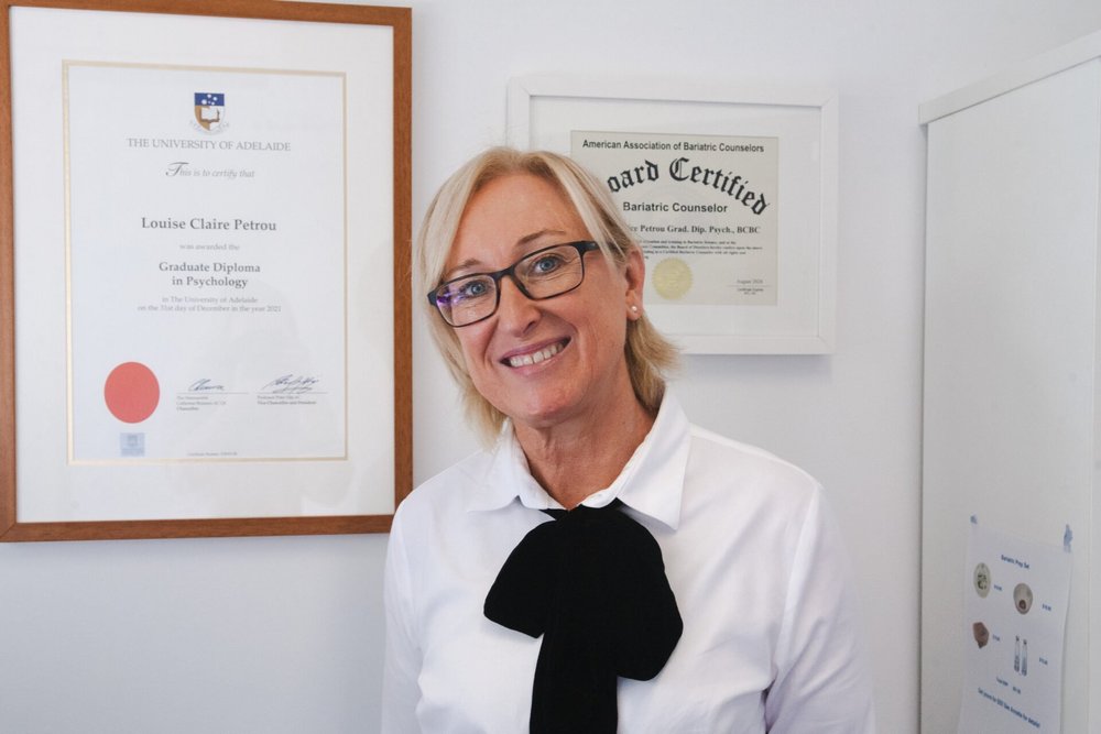 A woman with blonde hair, glasses, and a white shirt standing in front of framed diplomas on a white wall